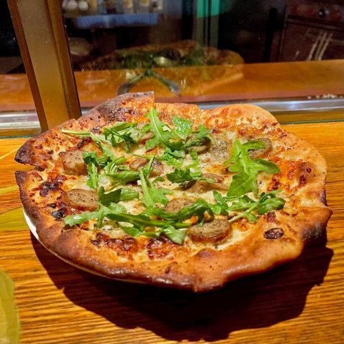 A freshly baked pizza topped with sausage, arugula, and melted cheese on a golden crust, served on a wooden table.