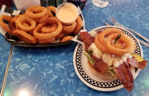 A plate with a burger topped with bacon and onion rings, alongside a large serving of crispy onion rings and dipping sauce.
