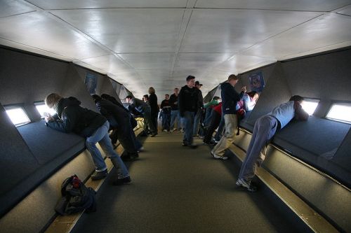 A group of people leaning against windows in a narrow, modern corridor, looking out at a view.