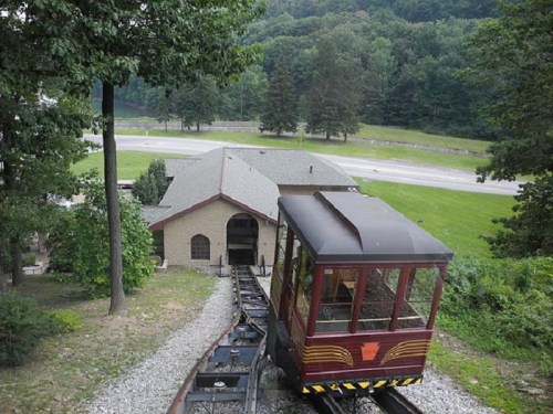 A vintage funicular railway car travels down a track towards a building, surrounded by green trees and hills.