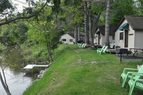 Cottages by a river, with green chairs and a dock, surrounded by trees and grass.