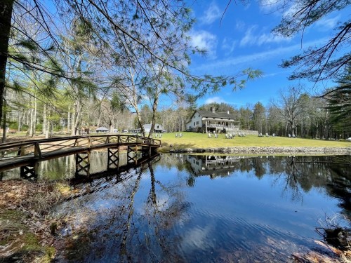 A serene landscape featuring a wooden bridge over a calm pond, with a house and trees in the background under a blue sky.