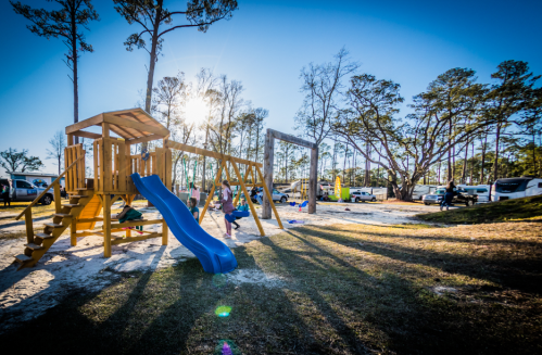 A playground with a blue slide and swings, surrounded by trees and RVs under a bright sun.