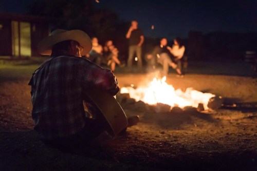 A person in a cowboy hat plays guitar by a campfire, surrounded by friends enjoying the night.