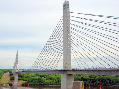 A modern cable-stayed bridge spans a river, featuring tall concrete towers and a backdrop of green trees.