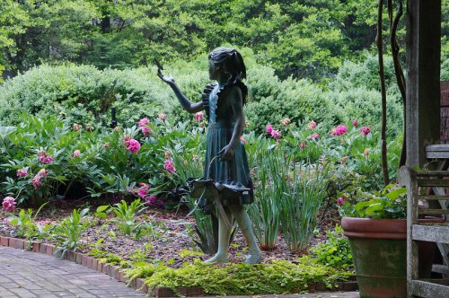 A bronze statue of a girl in a dress, reaching out to a butterfly, surrounded by colorful flowers and greenery.