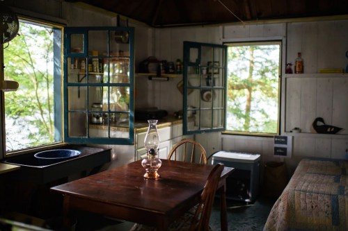 Cozy cabin interior with a wooden table, lantern, and blue-framed windows overlooking a natural landscape.