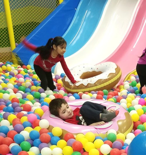 Two children play in a colorful ball pit; one slides down a slide while the other relaxes in a donut-shaped float.
