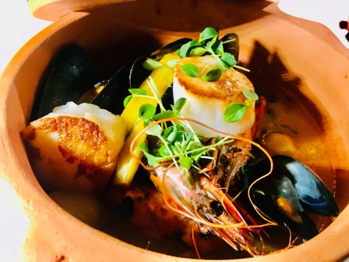 A clay pot filled with seafood, including shrimp, scallops, and mussels, garnished with fresh herbs.
