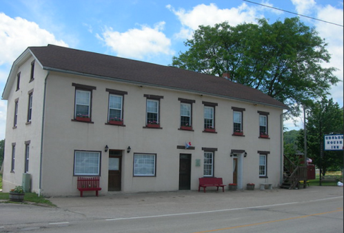 Historic two-story inn with red shutters and benches, surrounded by greenery under a partly cloudy sky.