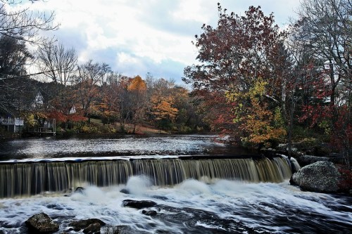A serene river scene with a waterfall, surrounded by autumn foliage and cloudy skies.