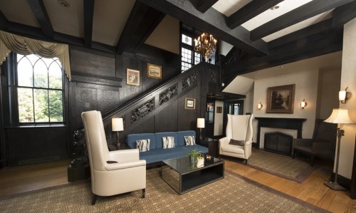 Elegant living room with dark wood beams, a staircase, two white chairs, a coffee table, and a cozy fireplace.