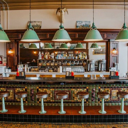 A vintage bar with green pendant lights, a wooden counter, and retro stools, featuring a well-stocked back shelf.