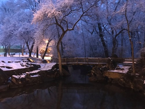 A serene winter scene featuring a snow-covered bridge over a calm stream, surrounded by frosty trees at dusk.