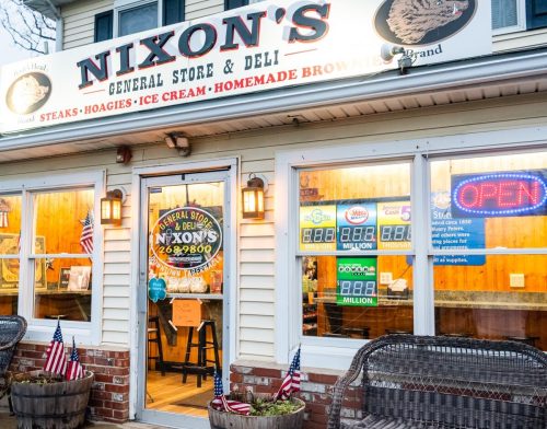 Exterior of Nixon's General Store & Deli, featuring a welcoming entrance with flags, benches, and a bright "Open" sign.