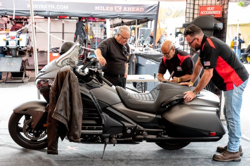 Three men inspect a motorcycle at a vendor booth, discussing its features and accessories.
