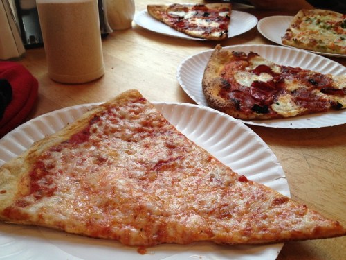 A slice of cheese pizza on a paper plate, with additional pizzas in the background.