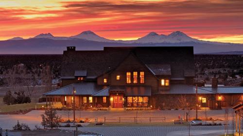 A large, rustic house with glowing windows against a vibrant sunset and snow-capped mountains in the background.