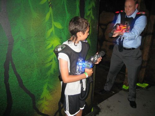 A child and an adult in vests hold laser tag guns in a colorful, jungle-themed environment.