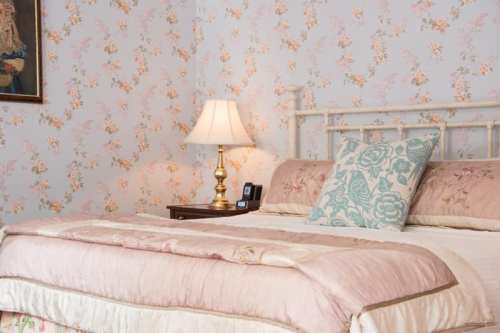 A cozy bedroom with floral wallpaper, a bed with decorative pillows, and a bedside lamp on a small table.