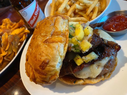 A close-up of a burger with grilled meat, pineapple salsa, and melted cheese, served with fries and a bottle of soda.