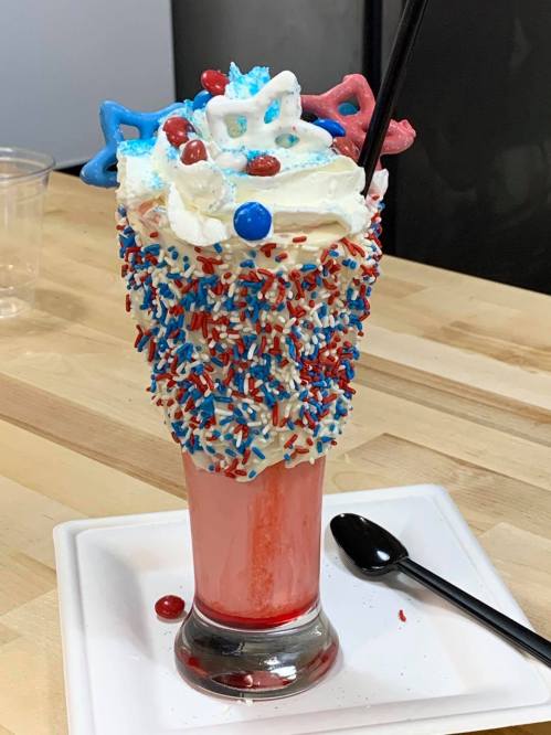 A tall milkshake topped with whipped cream, colorful sprinkles, and festive decorations in red, white, and blue.