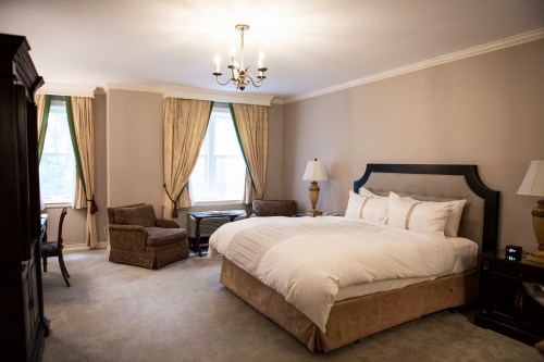 A spacious hotel room featuring a large bed, armchair, desk, and elegant curtains by the windows.
