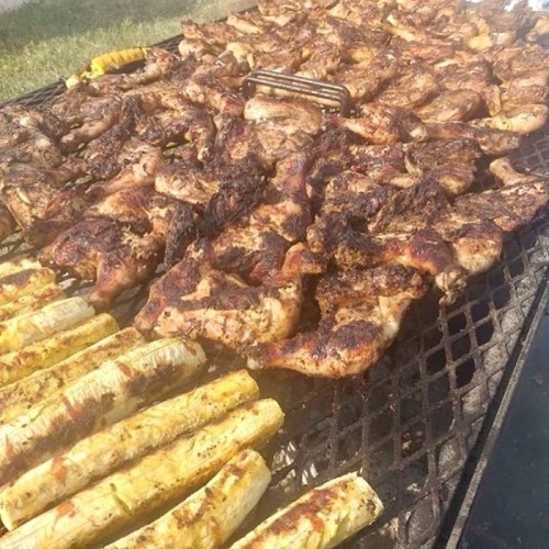 A large grill filled with marinated chicken pieces and grilled plantains, all sizzling in the sunlight.