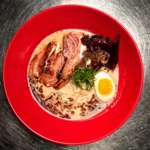 A vibrant red bowl of ramen featuring sliced meat, a soft-boiled egg, green onions, and dark leafy greens in rich broth.