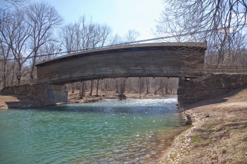 A wooden covered bridge spans a clear river, surrounded by bare trees and a serene landscape.