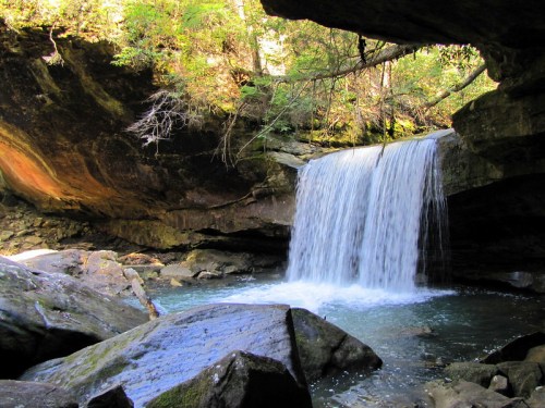 A serene waterfall cascades over rocks into a clear pool, surrounded by lush greenery and rugged cliffs.