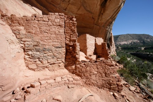 Ancient stone ruins nestled under a rocky overhang, surrounded by a scenic landscape of hills and trees.