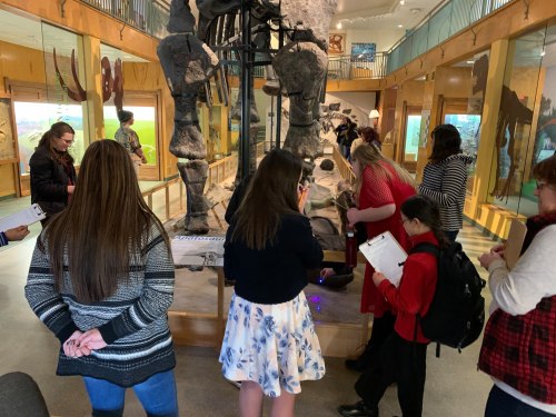 Visitors explore a museum exhibit featuring a large dinosaur skeleton, taking notes and interacting with displays.
