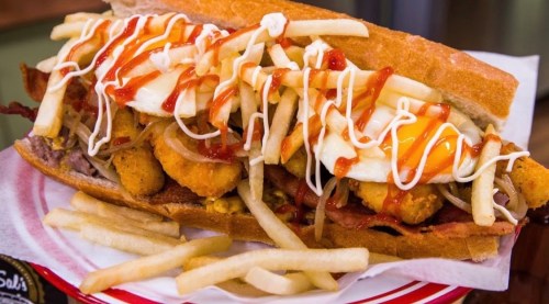 A large sandwich filled with fried items, bacon, fries, and drizzled with ketchup and mayonnaise.