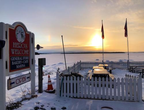 A snowy landscape with a sign for "Hero's Welcome" and a sunset over a frozen lake, featuring picnic tables and flags.