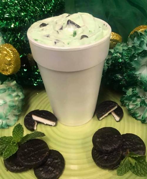 A white cup filled with mint green dessert topped with crushed cookies, surrounded by mint leaves and decorative green items.
