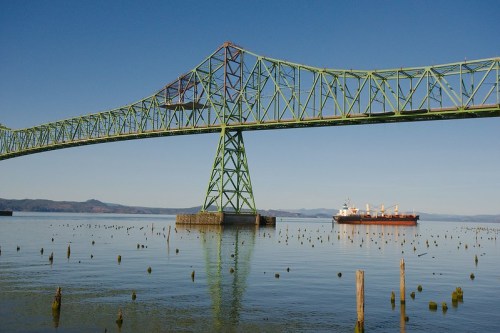 A green bridge spans over calm water, with a cargo ship passing beneath and wooden pilings visible in the foreground.