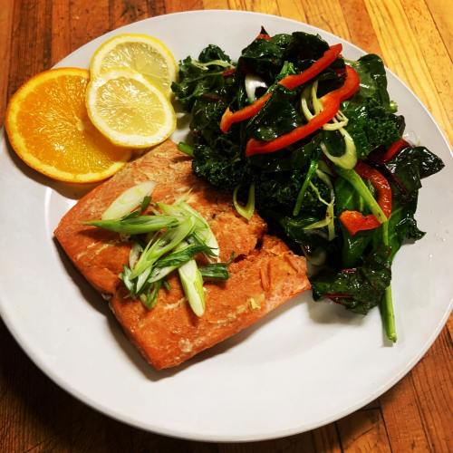 A plate featuring grilled salmon, sautéed greens with red peppers, and lemon slices.