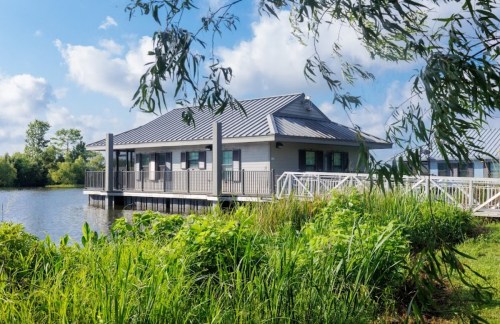 A modern building on stilts by a calm lake, surrounded by lush greenery and blue skies.