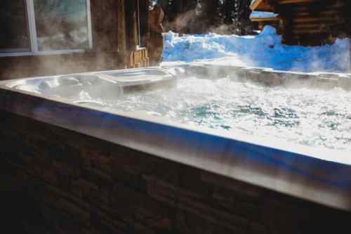 A steaming hot tub surrounded by snow, with a wooden cabin in the background.