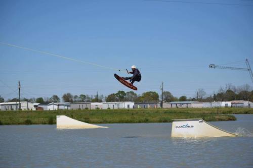 A person jumps on a wakeboard over a ramp in a lake, with a clear blue sky and buildings in the background.