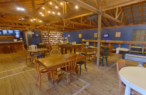Interior of a rustic restaurant with wooden tables, chairs, and a cozy atmosphere, featuring shelves and a bar area.