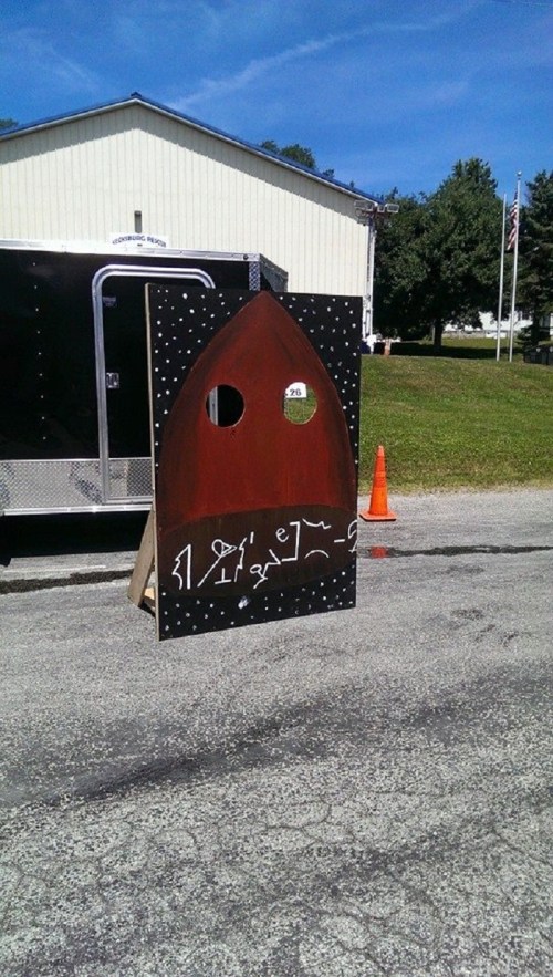 A wooden cutout shaped like a rocket, with a circular window, set outdoors near a trailer and grassy area.