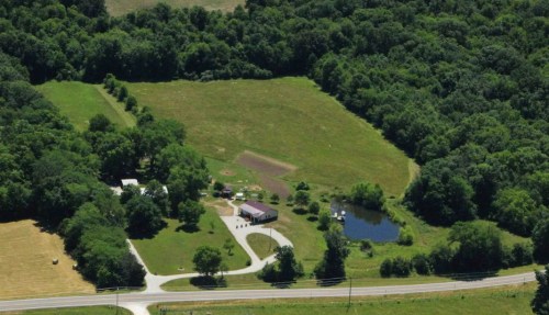 Aerial view of a rural property with a house, pond, and surrounding greenery, near a winding road.