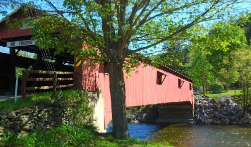 A red covered bridge spans a river, surrounded by greenery and a tree, with a sign reading "No Trucks."