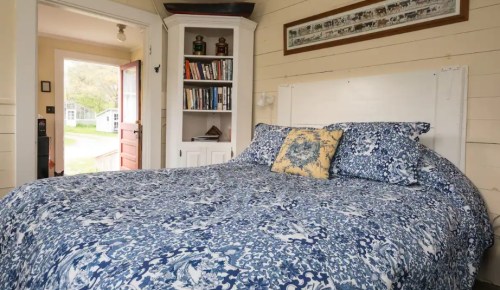 Cozy bedroom with a blue patterned bedspread, bookshelf, and an open door leading to a sunny outdoor view.