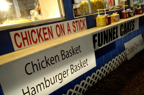 A food stand featuring signs for chicken on a stick, chicken basket, hamburger basket, and funnel cakes.