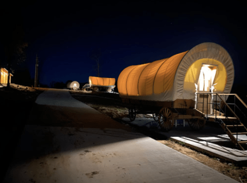 Three illuminated covered wagons on a pathway at night, surrounded by darkness and trees.