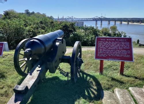 A historic cannon named "The Widow Blakely" with a river and bridge in the background, accompanied by an informational sign.