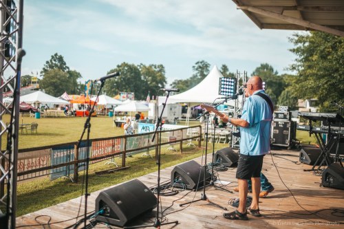 A musician performs on stage at an outdoor festival, with tents and a crowd visible in the background.
